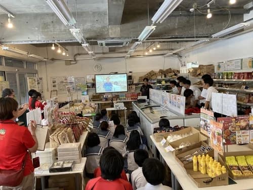 ecoeat 那覇与儀店の店内で、モニターの説明を聞く南風原中学校の生徒たちの後ろ姿