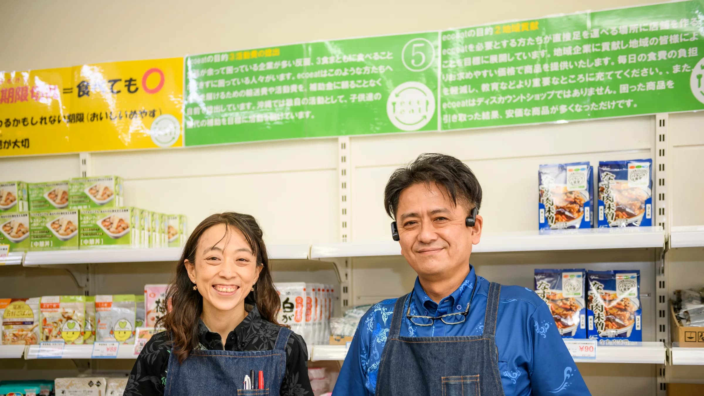 商品棚の前に並んで立つ玉城郁子さんと玉城淳一郎さん。上には「ecoeatはディスカウントショップではありません」などと書かれた掲示