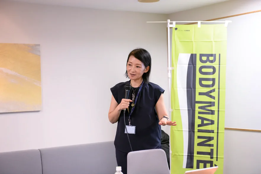 ボディメンテののぼりの横でマイクを持って話す葛西雅美氏